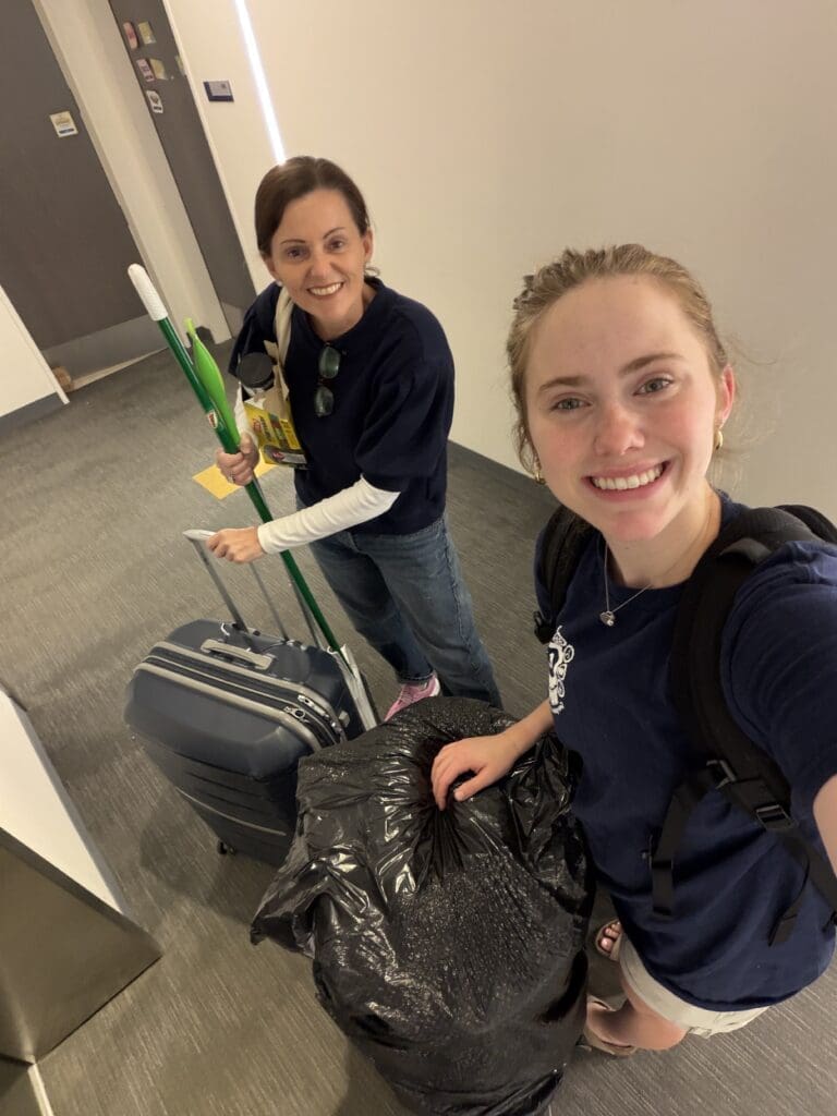 Two women wearing navy shirts stand in a hallway, smiling at the camera. One holds a suitcase and cleaning supplies—perfect for college move out organization—while the other carries a large trash bag and wears a backpack. The setting appears to be indoors, possibly a dorm.