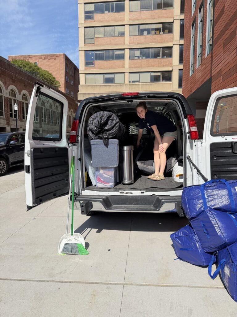 A person stands in the back of an open van filled with packed bins, bags, and household items, showing smart college move out organization. A broom and blue bags are on the ground next to the van, parked near tall buildings on a sunny day.