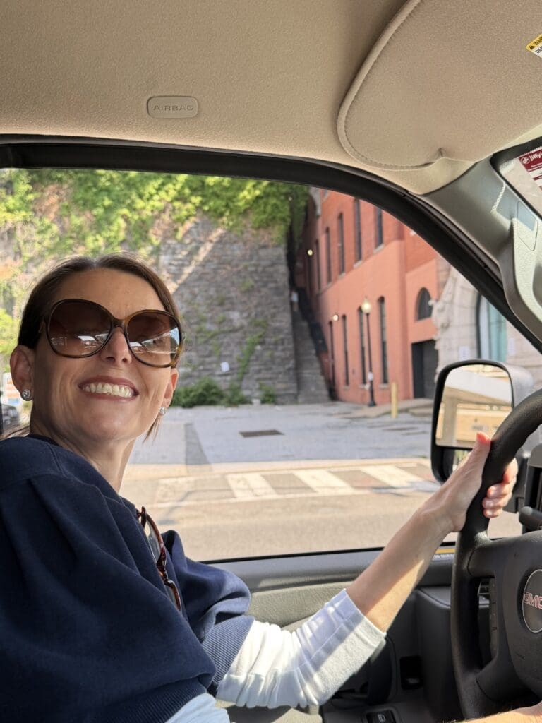 A woman wearing sunglasses and smiling sits in the driver’s seat, ready for a college move out organization day. Sunlight shines in, with a brick building and stone staircase visible through the window outside.