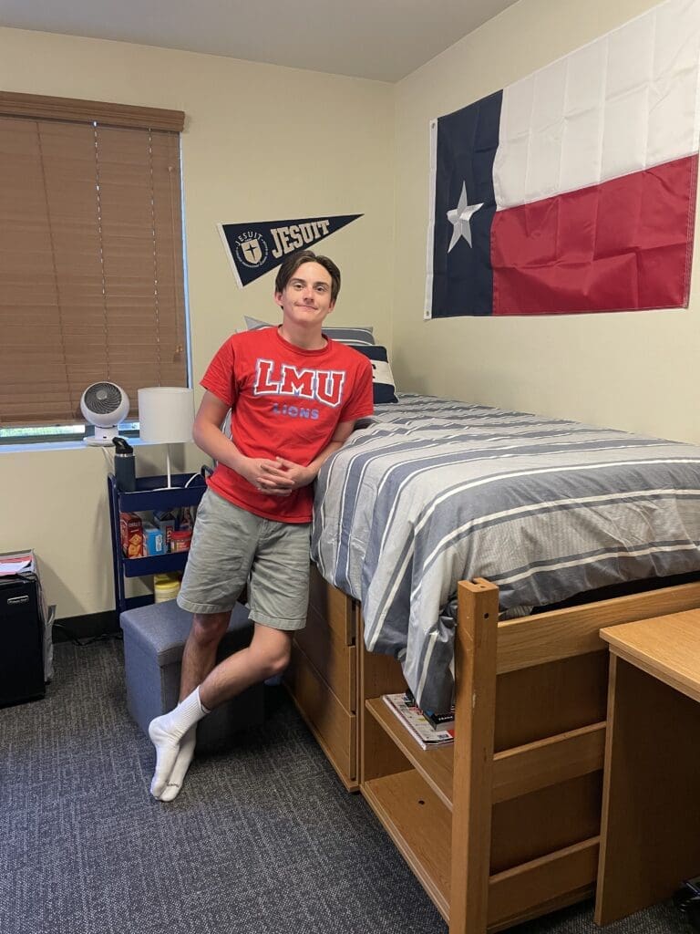 A young man in a red LMU Lions shirt and gray shorts leans against a wooden bed in a dorm room, surrounded by college move out organization essentials. Behind him are a Texas flag, a Jesuit pennant, and a bookshelf with various items.