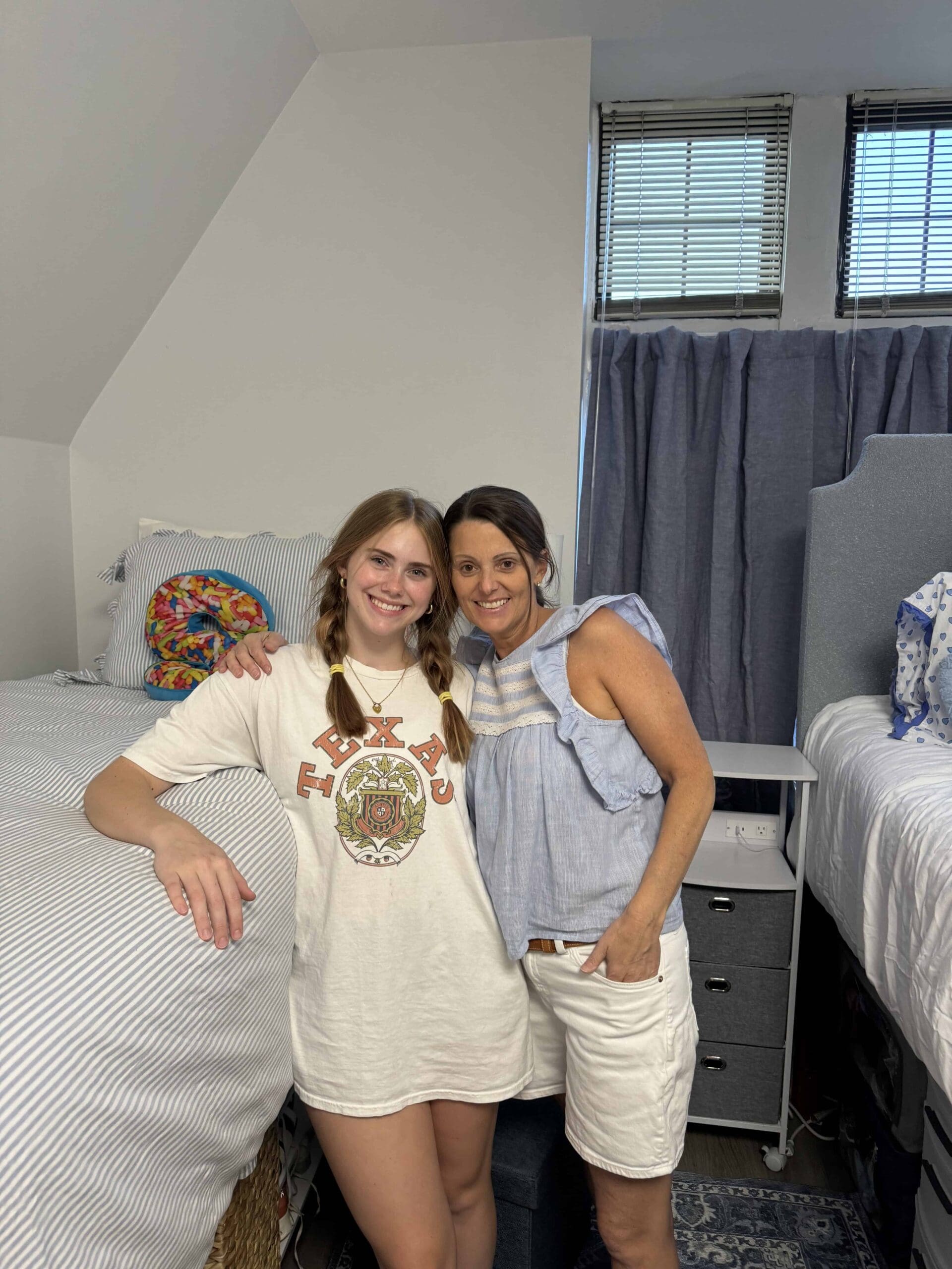 A mom and her college student daughter smile and stand arm-in-arm in a bright dorm room with neatly made beds, light bedding, and a window with blinds and curtains in the background.