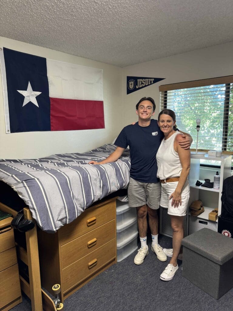 A young college student and his mom smile and stand together in a dorm room with a Texas flag on the wall, a neatly made bed, and a window letting in natural light.