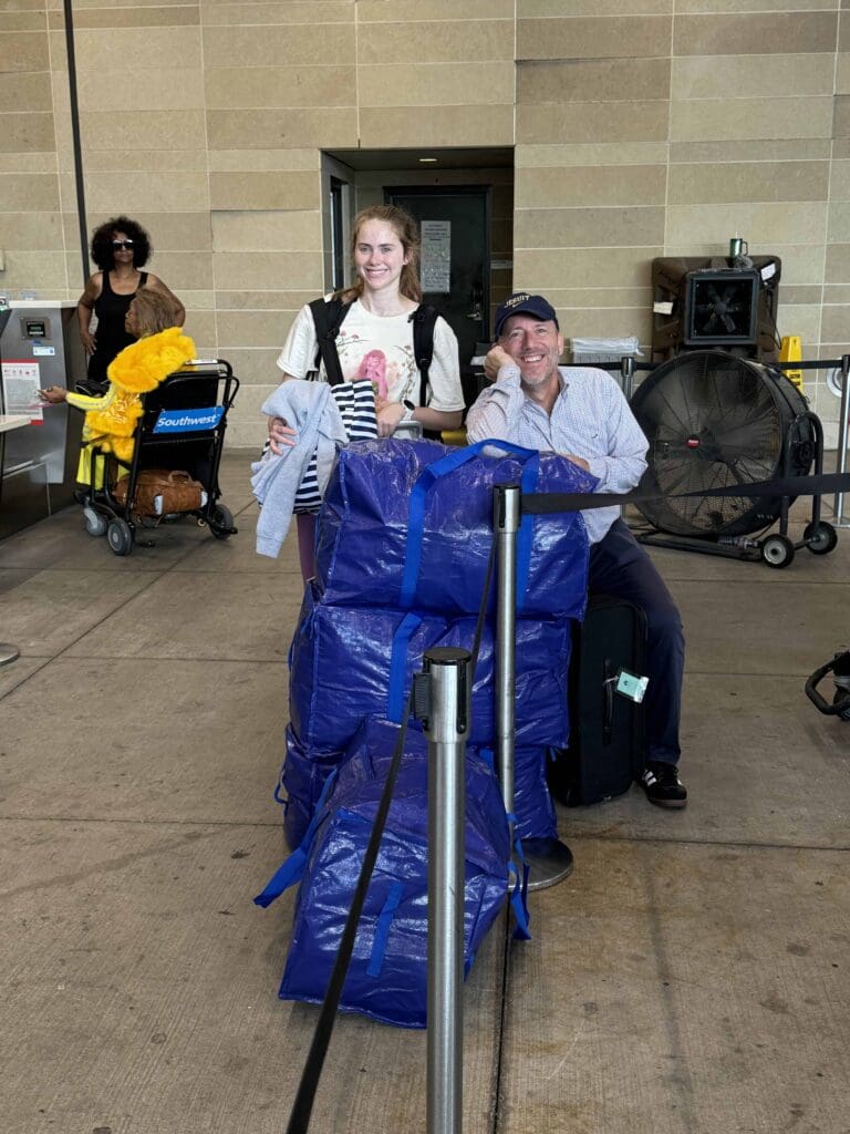 A smiling young college student and her dad stand with blue bags at an airport curbside. Behind them, a woman pushes a yellow plush toy in a stroller. A large fan and brick wall are visible in the background.