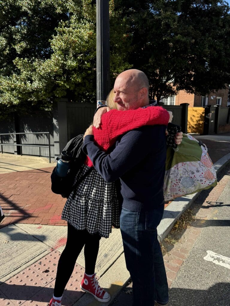 A college student and her dad hug warmly on a sunny sidewalk. The woman wears a red sweater, black checkered skirt, and red sneakers. The man is in a navy sweater and jeans. Trees and a fence are in the background.