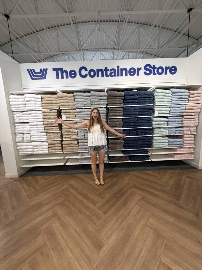 A college student stands with arms outstretched in front of neatly stacked towels in various colors at The Container Store.