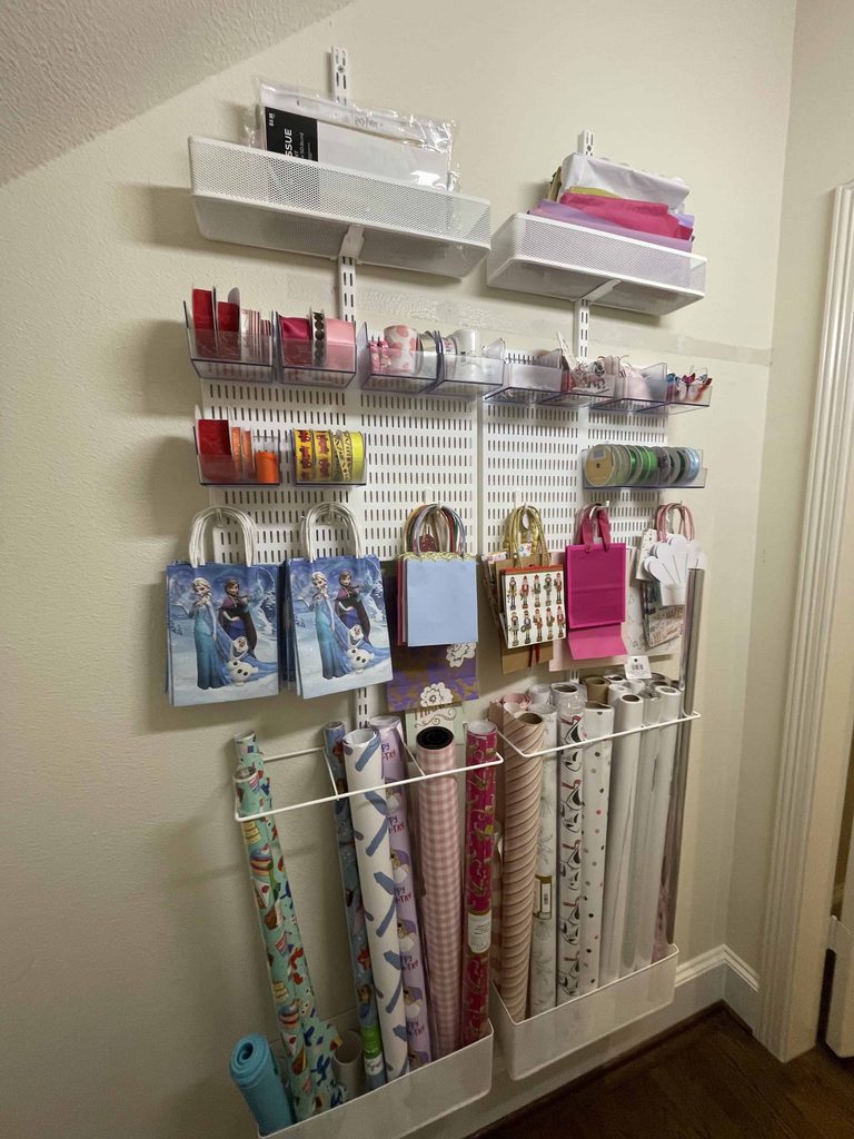 Wall-mounted organizer holds ribbons, gift bags, and wrapping paper rolls on shelves and racks, neatly arranged in a corner of a room with neutral walls.