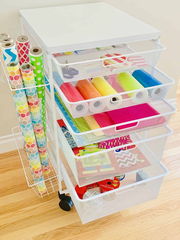 A white rolling cart with mesh drawers holds colorful tissue paper, gift bags, and other wrapping supplies. Rolls of wrapping paper stand upright in a side holder. The cart is on a wooden floor next to a beige wall.