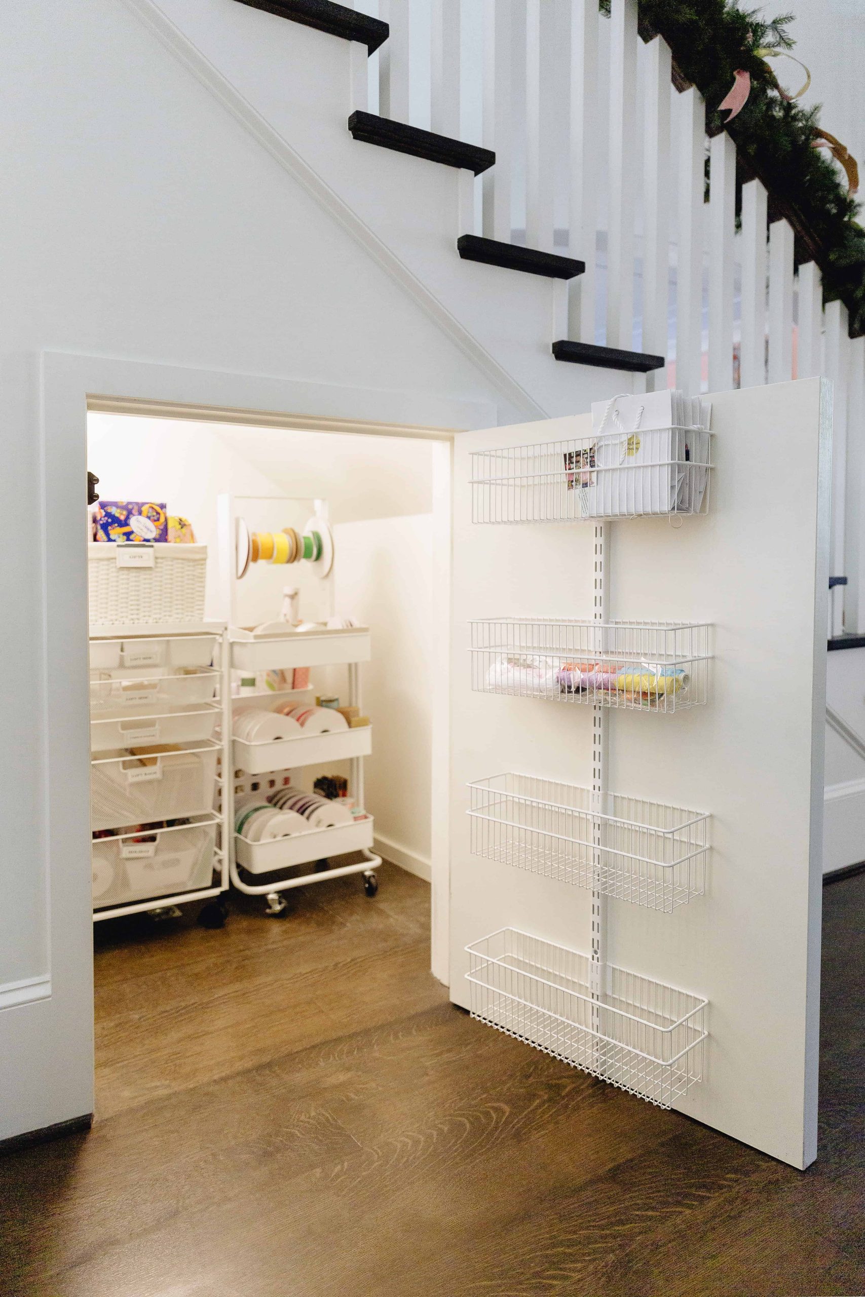 A small storage room under stairs with white shelves, stacked bins, and baskets on the door. Various craft supplies, ribbons, and organization boxes are neatly arranged in a gifting closet in Dallas. The floor is dark wood.