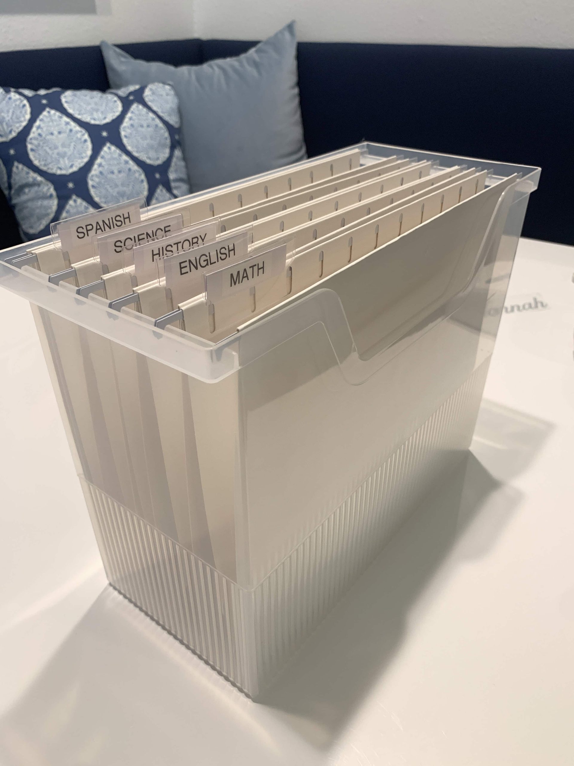 Clear ribbed plastic file box on a white table, holding cream hanging folders with clear tabs labeled Math, English, History, Science, and Spanish.