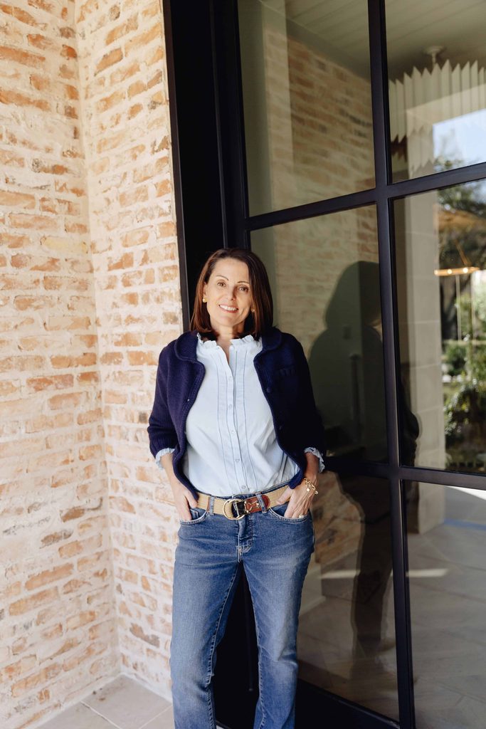 Jenny Dietsch, owner of Getting It Done Organizing, in a light blue ruffled blouse, navy cardigan, and jeans, leaning against a brick wall beside a black-framed glass door.