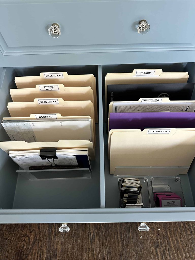Open light blue drawer with two sections of upright file folders on acrylic stands, labeled Bills to Pay, Things to Do, 2025 Taxes, Banking, Notary, Manuals & Gadgets, and To Shred, plus small clear bins for cards and a notary stamp.